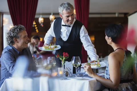 Ein lächelnder Kellner serviert einem reifen Paar am Tisch im Restaurant ausgefallene Gerichte, lizenzfreies Stockfoto