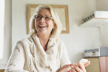Lächelnde ältere Frau im Pullover, die in der Küche mit ihrem Handy SMS schreibt - CAIF15899