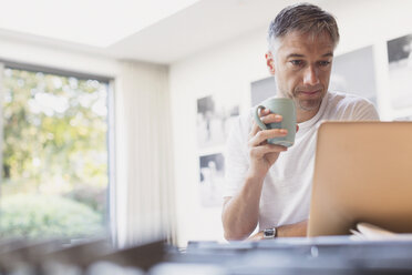 Mann trinkt Kaffee und arbeitet am Laptop in der Küche - CAIF15955