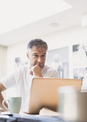 Mann trinkt Kaffee und arbeitet am Laptop in der Küche - CAIF15958