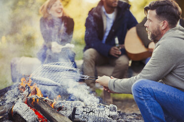 Man frying fish in grill basket over campfire near friends - CAIF15994