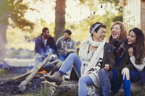 Female friends laughing and roasting marshmallows at campfire - CAIF16003