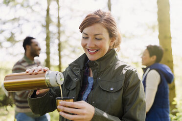 Smiling woman pouring coffee from insulated drink container in woods - CAIF16004