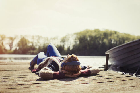Woman relaxing laying on dock listening to music with headphones at sunny lakeside - CAIF16006