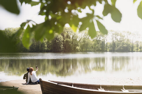 Couple with camera phone taking selfie on sunny lakeside dock next to canoe - CAIF16008