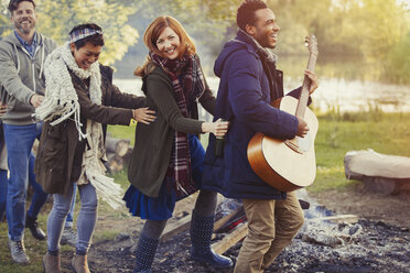 Playful friends with guitar dancing in conga line at campsite - CAIF16017