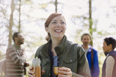 Lächelnde Frau trinkt Kaffee aus einem isolierten Getränkebehälter beim Wandern im Wald - CAIF16030