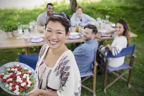 Porträt einer lächelnden Frau, die ihren Freunden am Tisch einer Gartenparty Caprese-Salat serviert - CAIF16060