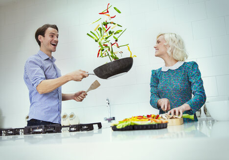Überraschtes Paar beim Kochen und Wenden von Gemüse in der Bratpfanne in der Küche - CAIF16101