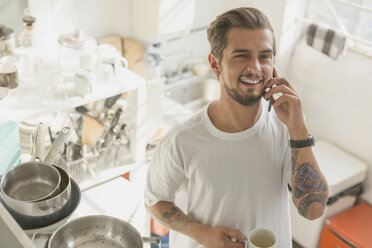 Tätowierter junger Mann, der in der Wohnküche mit seinem Handy telefoniert und Kaffee trinkt - CAIF16142