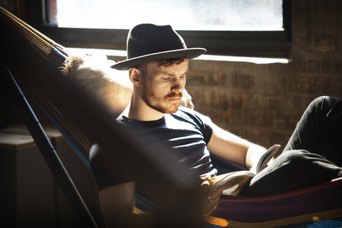 Mann mit Hut, der ein Buch liest, während er sich in der Hängematte entspannt, lizenzfreies Stockfoto