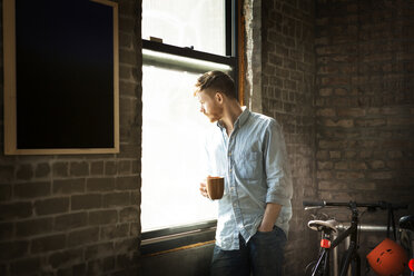 Mann mit Hand in der Tasche, der eine Kaffeetasse hält, während er zu Hause am Fenster steht - CAVF07934