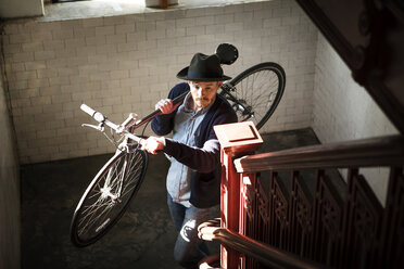 Hoher Blickwinkel eines Mannes, der ein Fahrrad auf einer Treppe zu Hause trägt - CAVF07940
