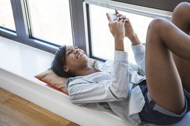 Hochformatige Ansicht einer Frau, die ein Smartphone benutzt, während sie zu Hause am Fenster liegt - CAVF07957