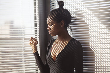 Woman looking down while standing by blinds at home - CAVF07964