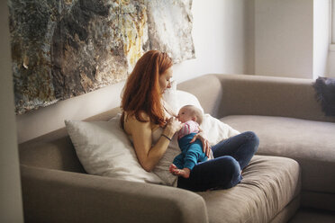 Mother feeding baby girl while sitting on sofa at home - CAVF07974