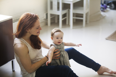 Mother holding daughter while sitting on floor at home stock photo