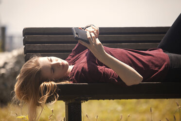 Woman using mobile phone while lying on bench at park - CAVF08127