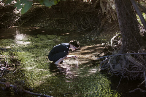 Side view of teenage girl holding water while crouching in stream - CAVF08185