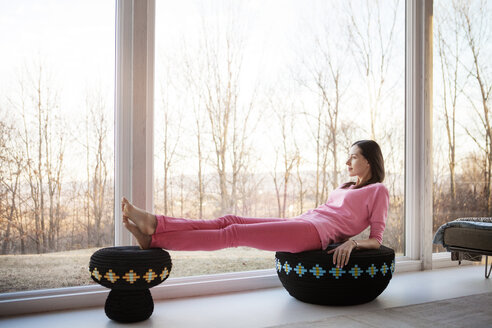 Seitenansicht einer Frau, die sich zu Hause auf einem Hocker am Fenster entspannt - CAVF08254