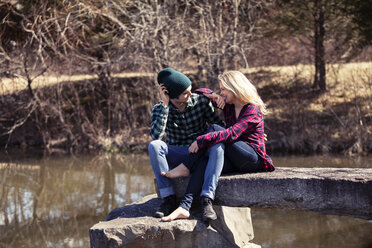 Happy couple talking while sitting on rock by lake - CAVF08264