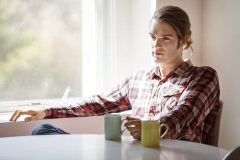Nachdenklicher Mann hält Kaffeetasse, während er zu Hause am Fenster sitzt, lizenzfreies Stockfoto