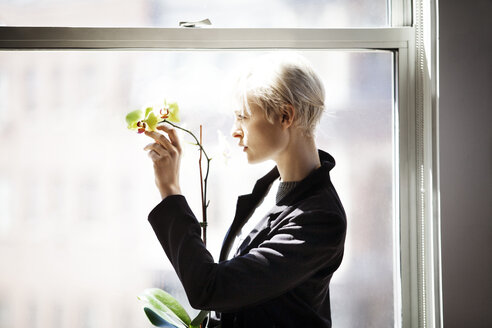 Seitenansicht einer nachdenklichen Frau, die Blumen hält, während sie durch ein Fenster zu Hause schaut - CAVF08285