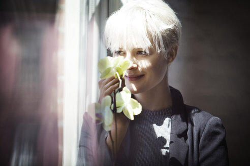 Nachdenkliche Frau mit Blumen in der Hand und Blick durch das Fenster zu Hause - CAVF08286