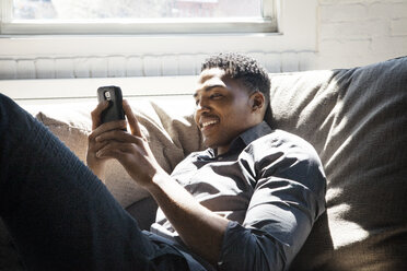 Smiling man using smart phone while lying on sofa at home - CAVF08299