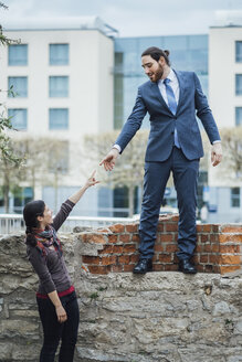 Businessman standing on a wall reaching out his hand for woman - JSCF00070
