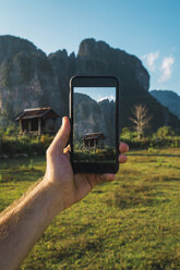 Laos, hand holding smartphone, Display with wooden hut and mountains - KKAF00911
