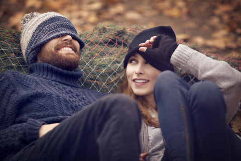 Frau sieht Mann mit Strickmütze an, der sich an den Weihnachtsbaum lehnt, lizenzfreies Stockfoto