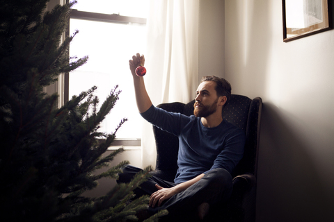 Mann betrachtet Weihnachtsschmuck, während er zu Hause auf einem Sessel am Weihnachtsbaum sitzt, lizenzfreies Stockfoto