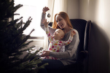 Mutter mit Tochter, die mit Weihnachtsschmuck spielt, während sie zu Hause auf einem Sessel sitzt - CAVF08363