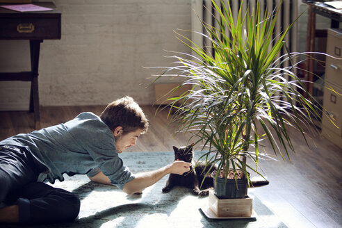 Man playing with cat while lying on floor at home - CAVF08391