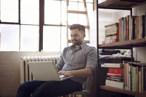 Mann benutzt einen Laptop, während er zu Hause auf einem Stuhl sitzt, lizenzfreies Stockfoto