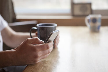 Ausgeschnittenes Bild eines Mannes, der ein Mobiltelefon benutzt und eine Kaffeetasse in einem Café hält - CAVF08501