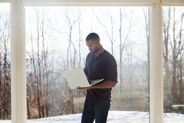 Mann benutzt Laptop, während er zu Hause vor einem Glasfenster steht - CAVF08541