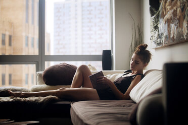 Woman reading book while sitting on sofa at home - CAVF08605