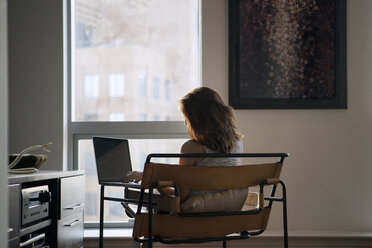 Rear view of woman using laptop while sitting on chair at home - CAVF08625