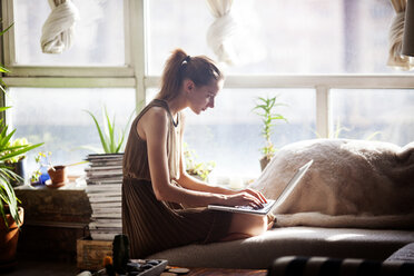 Frau, die einen Laptop benutzt, während sie zu Hause auf dem Sofa am Fenster kniet - CAVF08676
