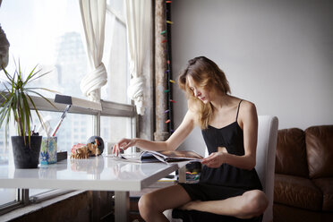 Frau liest eine Zeitschrift, während sie auf einem Stuhl am Tisch zu Hause sitzt - CAVF08678