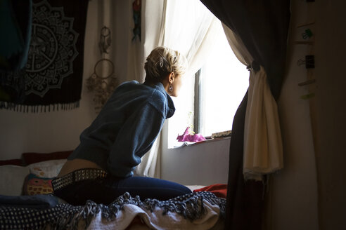 Seitenansicht einer Frau, die durch das Fenster schaut, während sie zu Hause auf dem Bett sitzt - CAVF08695
