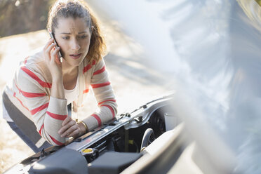 Frustrierte Frau, die mit ihrem Handy telefoniert und sich den Motor ihres Autos am Straßenrand ansieht - CAIF16948