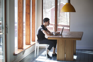 Seitenansicht eines Mannes, der einen Laptop benutzt, während er zu Hause auf einem Stuhl am Tisch sitzt - CAVF08772