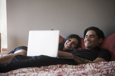 Lächelndes Paar, das einen Laptop benutzt, während es zu Hause auf dem Bett liegt - CAVF08775