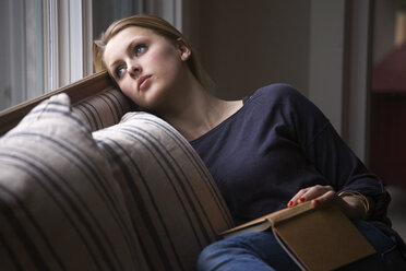Nachdenkliches Mädchen mit Buch in der Hand, das wegschaut, während es sich auf dem Sofa zu Hause entspannt - CAVF08848