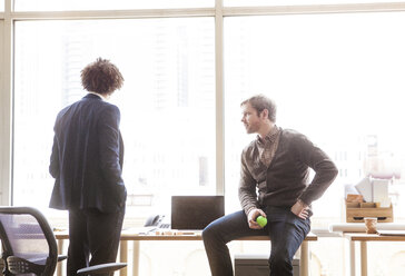 Businessman looking at coworker while sitting on table at office - CAVF08931