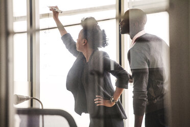 Geschäftsfrau, die an eine Glaswand schreibt, während sie neben einem Kollegen im Büro steht - CAVF08933