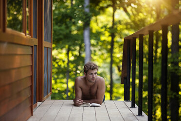 Junge mit Buch, der wegschaut, während er auf dem Balkon auf einer Diele liegt - CAVF09126